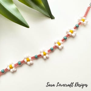 May include: A handmade bracelet featuring white daisy-shaped beads with yellow centers, interspersed with turquoise beads. The bracelet is strung on a pink cord. The text "Sara Lovecraft Designs" is at the bottom.