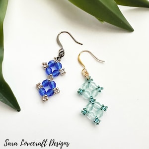 May include: Two pairs of handmade beaded earrings. One pair features dark blue beads with silver accents and black ear wires. The other pair has light blue beads with gold ear wires. The earrings are displayed on a white surface with green leaves.