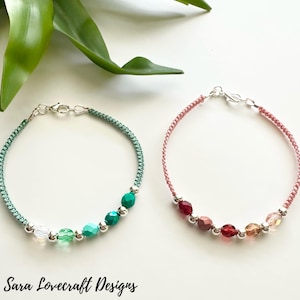 May include: Two handmade bracelets with colorful beads. One bracelet is teal with green and clear beads, the other is pink with red, pink, and amber beads. Both have silver clasps and are on a white surface.