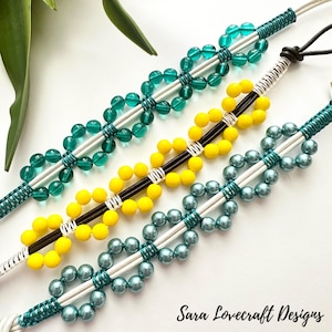 May include: Three beaded bracelets in teal, yellow, and teal-blue. Each bracelet features a central design of connected beads, with a contrasting color cord. The bracelets are displayed on a white surface. The text "Sara Lovecraft Designs" is visible.