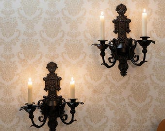May include: Two black metal wall sconces, each featuring an ornate backplate and two curved arms holding lit white taper candles. The sconces are mounted symmetrically, showcasing intricate scrollwork and floral-inspired decorative details.