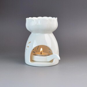 Puede incluir: Un quemador de aceite de cerámica blanca con un cuenco superior festoneado y un diseño recortado en el cuerpo. Una pequeña vela de té con una llama encendida se encuentra dentro de la abertura arqueada sobre una pequeña bandeja adjunta.