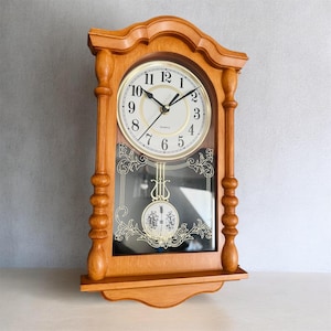 May include: A wooden wall clock with a light brown finish, featuring a round white face with black Arabic numerals and black hands. Below the clock face is a glass panel with gold decorative filigree and a gold-coloured pendulum inside.