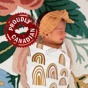 May include: A white swaddle blanket featuring a rainbow pattern in shades of brown, pink, and yellow. The blanket has the name "Emmy" printed on it three times. The blanket is wrapped around a baby wearing a brown headband.