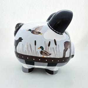 May include: A ceramic piggy bank with a hand-painted design featuring ducks, a deer, and a black dog in a marsh scene. The ears are black, and the base has a black and white checkered pattern. The bank is a decorative item.