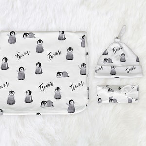 May include: A baby gift set with a white blanket, hat, and headband featuring a black and white penguin pattern and the name "Trevor" repeated throughout.