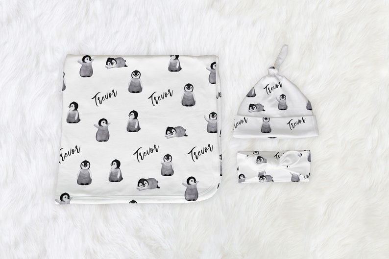 May include: A baby gift set with a white blanket, hat, and headband featuring a black and white penguin pattern and the name "Trevor" repeated throughout.