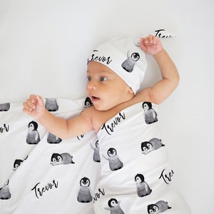 May include: A white swaddle blanket and matching hat with a penguin pattern and the name "Trevor" printed on them. A baby is lying on the blanket.