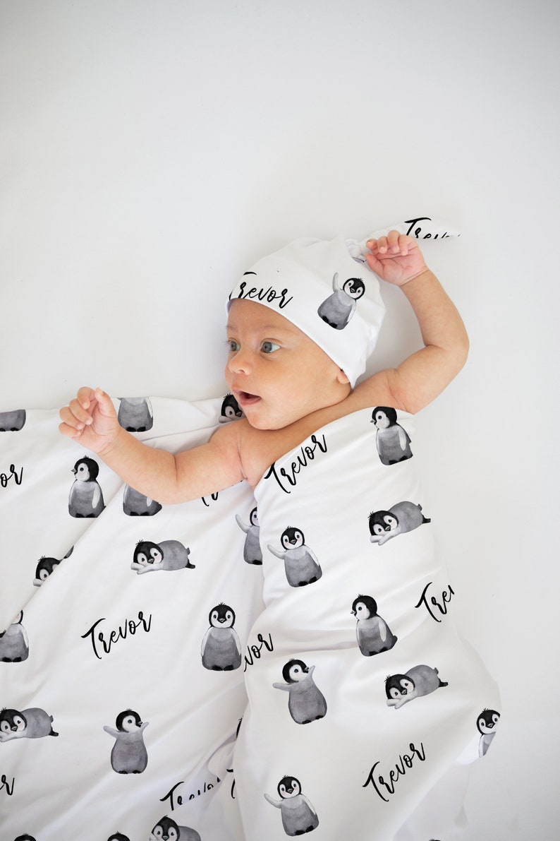 May include: A white swaddle blanket and matching hat with a penguin pattern and the name "Trevor" printed on them. A baby is lying on the blanket.