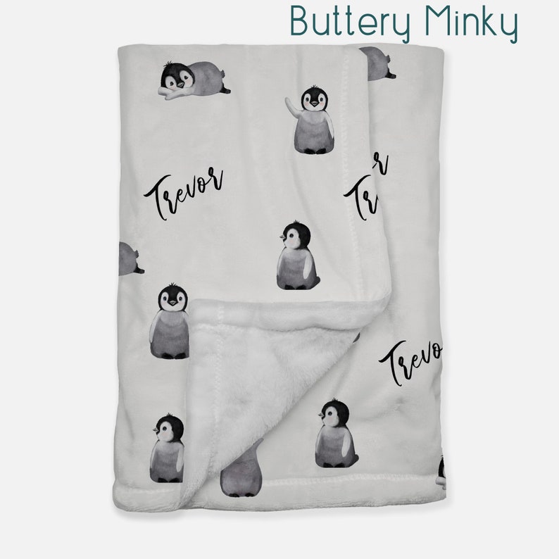May include: A white and gray baby blanket with a buttery minky texture. The blanket features a repeating pattern of black and white penguins. The text "Trevor" is written in black cursive on the blanket.
