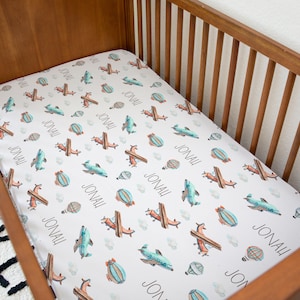 Pode incluir: Um lençol de berço branco com um padrão de avião e balão de ar quente azul e laranja. O lençol é personalizado com o nome "JONALI" repetido em todo o design.