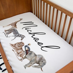 May include: White crib sheet with a safari animal print design. The sheet features a giraffe, zebra, lion, elephant, and a bird. The name "Michael" is written in cursive across the sheet.