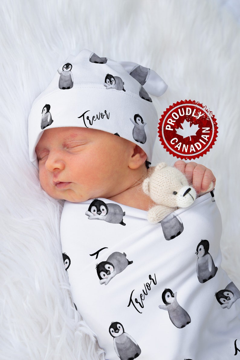 May include: A white swaddle blanket with a penguin pattern and the name "Trevor" written in black. A baby is sleeping in the swaddle, wearing a white penguin hat with the name "Trevor" written in black. A small white teddy bear is in the baby's hand. A red and white sticker with the text "Proudly Canadian" is in the background.