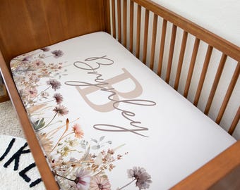 Puede incluir: Una sábana bajera para cuna con un diseño floral y el nombre "Brynley" en letra cursiva. La sábana tiene un fondo crema con flores de acuarela en tonos rosa, marrón y verde. La cuna es de madera.