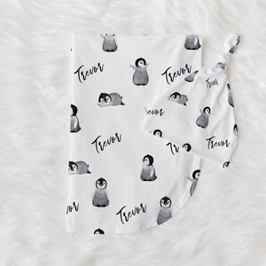 May include: White baby blanket with a penguin pattern and the name "Trevor" repeated throughout. The blanket is paired with a matching burp cloth.