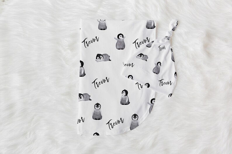 May include: White baby blanket with a penguin pattern and the name "Trevor" repeated throughout. The blanket is paired with a matching burp cloth.