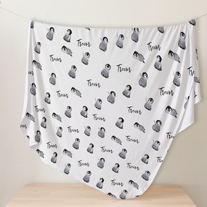 May include: White baby blanket with a repeating pattern of black and white penguins. The name "Trevor" is printed on the blanket.