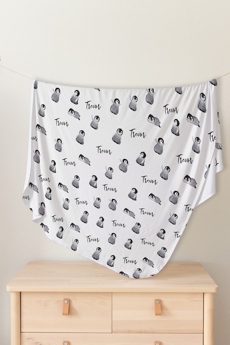 May include: White baby blanket with a repeating pattern of black and white penguins. The name "Trevor" is printed on the blanket.