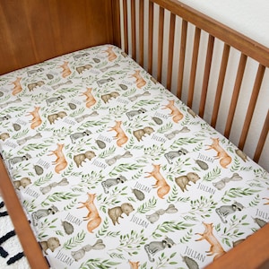Peut inclure: Un drap-housse pour lit bébé avec un motif d'animaux de la forêt. Le drap blanc présente des illustrations de renards, d'ours, de ratons laveurs et d'os, avec le nom "Julian" en écriture cursive. Des feuilles vertes sont dispersées.