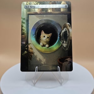 May include: A rectangular trading card featuring a holographic finish and the text Sol Ring. The illustration depicts a small orange and white kitten sitting inside the open circular door of a white washing machine, with green plants visible.