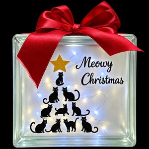 May include: A square glass block light featuring a red satin bow on top. The front displays a gold star and black silhouettes of cats arranged in a tree shape, with the text Meowy Christmas in black script. Internal white fairy lights glow.