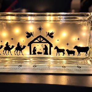May include: A rectangular glass block light featuring a black silhouette of a nativity scene with three wise men on camels, a stable, angels, and animals. The block is illuminated from within by warm lights and sits on a mat marked in inches.