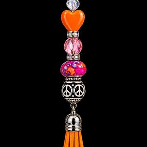 May include: A decorative bag charm featuring a silver-tone lobster clasp, a clear faceted bead, an orange heart-shaped bead, two crystal-studded spacers, a pink faceted bead, a multi-colored glass bead, and a silver-tone peace sign bead with an orange tassel.