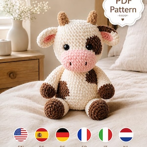 Puede incluir: Un peluche de vaca tejido a ganchillo con cuerpo color crema, manchas marrón oscuro, hocico rosa claro y pequeños cuernos marrones. El juguete tiene grandes ojos de seguridad negros y orejas rosas. Una etiqueta festoneada muestra el texto PDF Pattern.