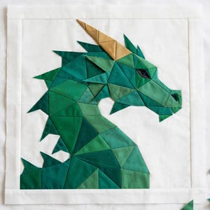 May include: A square fabric quilt block featuring a geometric dragon head design. The dragon is constructed from various shades of green fabric triangles with a single gold-colored horn and a dark eye, set against a plain white background.