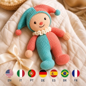 May include: A knitted jester doll featuring a two-toned colour scheme of coral pink and teal. The doll has a round beige face with black embroidered eyes and a small nose, a white ruffled collar, and a pointed hat with small pom-poms at the tips.