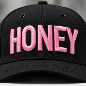 May include: A black baseball cap featuring the word HONEY embroidered in raised, light pink lettering across the front panels. The cap includes a structured crown, a curved brim, and dark metal eyelets for ventilation on the upper panels.
