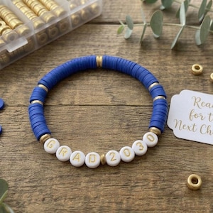 Può includere: Un braccialetto circolare composto da perline blu a forma di disco e distanziatori dorati. Il braccialetto presenta perline bianche rotonde con lettere dorate che compongono la scritta GRAD 2030. Un'etichetta rettangolare bianca con bordi smerlati mostra il testo Ready for the Next Chapter in corsivo dorato, accompagnato da un contenitore di plastica riempito con perline blu e dorate extra.