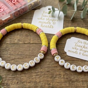May include: Two stretch bracelets made of yellow and pink clay disc beads with gold-tone spacers. Each bracelet features white round beads with gold lettering spelling out TEACHER. Included are white tags with the text For the Teacher who shapes minds & hearts.