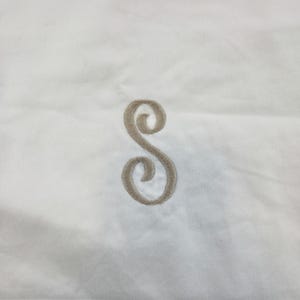May include: A white fabric surface featuring a centred, embroidered letter S in a light brown, cursive script font. The stitching is visible, creating a textured appearance for the monogram against the smooth, plain white textile background.