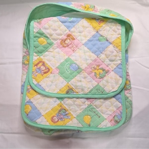 May include: A quilted fabric bag featuring a diamond pattern in pastel yellow, blue, pink, and green. The fabric is printed with Cabbage Patch Kids characters and balloons. The bag has a mint green trim and a flap closure with a soft texture.