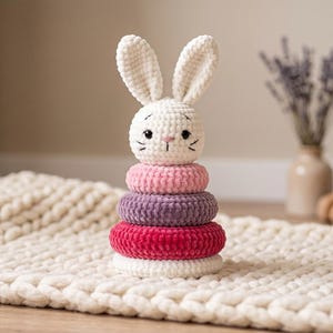 May include: A crochet stacking toy featuring a white rabbit head with long ears, black eyes, and embroidered whiskers. The base consists of four plush, circular rings in stacked colors of light pink, purple, dark pink, and white, made of soft yarn.
