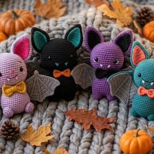 May include: Four crocheted bat plushies in light purple, black, dark purple, and teal. Each bat features large ears, grey wings, black bead eyes, small white fangs, and a small bow tie. The bats are arranged in a row with small orange pumpkins.