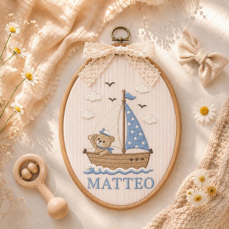 May include: An oval embroidery hoop featuring a stitched scene of a brown teddy bear in a sailboat on blue waves. The boat has a white sail and a blue polka-dot sail. Above are clouds and birds. The name MATTEO is embroidered in blue at the bottom.