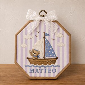 May include: An octagonal wooden embroidery hoop frame containing fabric with a light purple and white striped pattern. The embroidery features a brown teddy bear in a sailboat with white and blue polka dot sails, white clouds, birds, and the name MATTEO.