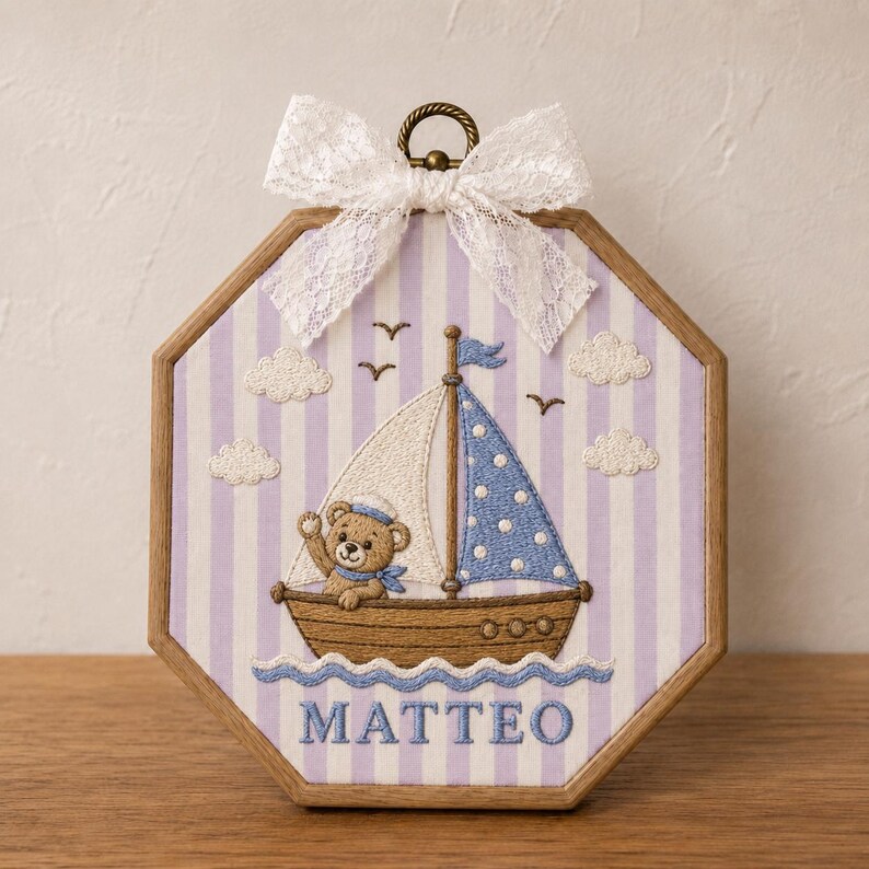 May include: An octagonal wooden embroidery hoop frame containing fabric with a light purple and white striped pattern. The embroidery features a brown teddy bear in a sailboat with white and blue polka dot sails, white clouds, birds, and the name MATTEO.