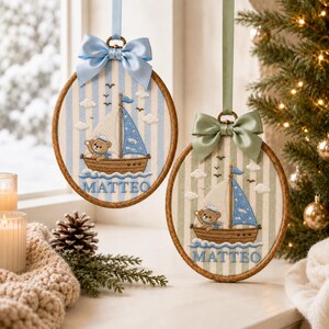 May include: Two oval embroidery hoops featuring a stitched sailboat with a teddy bear captain on a striped background. Each hoop displays the name MATTEO in blue thread and is topped with a satin ribbon bow in light blue or sage green.
