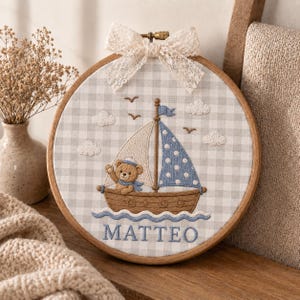 May include: A round wooden embroidery hoop featuring a light gray and white gingham fabric background. The embroidery depicts a brown teddy bear in a brown sailboat with a blue polka dot sail, white clouds, birds, and the name MATTEO in blue.