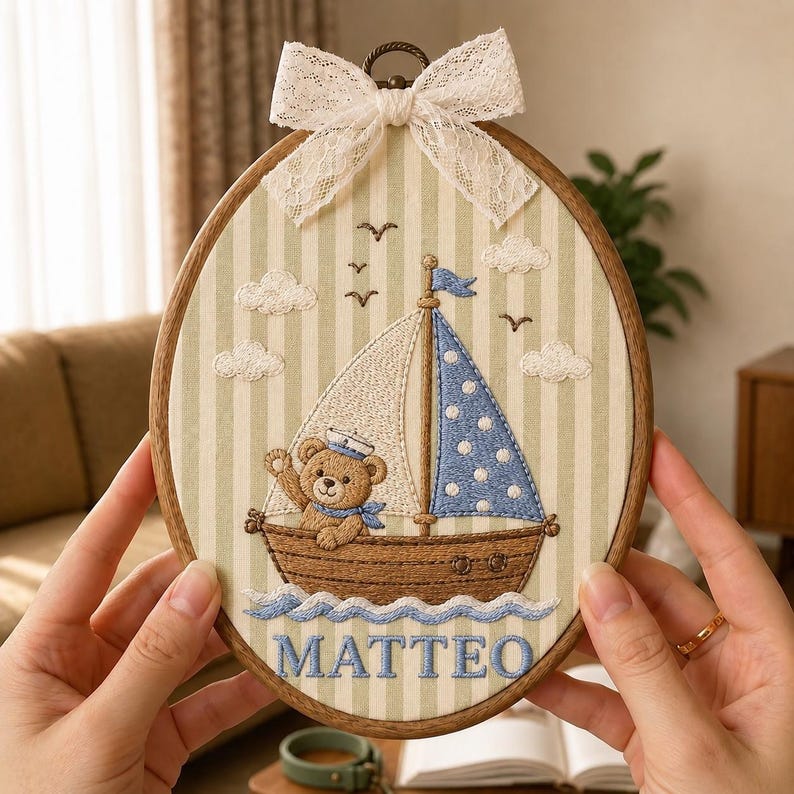 May include: An oval embroidery hoop featuring a brown teddy bear wearing a sailor hat inside a brown boat with a white and blue polka-dot sail. The background is light green and white striped fabric with embroidered clouds and birds, topped with a white lace bow and the name MATTEO in blue.