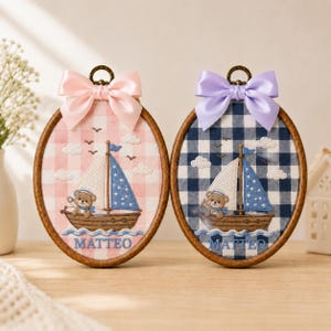 May include: Two oval embroidery hoops featuring a sailboat with a teddy bear captain. One hoop has a pink gingham background with a pink bow, the other a navy blue gingham background with a lavender bow. Both display the name MATTEO in blue text.