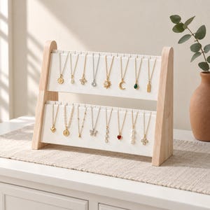 May include: A two-tiered wooden jewelry display stand with a light-colored frame and white fabric panels. The stand features multiple hooks holding various gold-toned necklaces with diverse pendants, including a sun, moon, heart, and pearls.