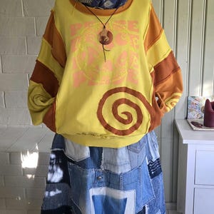 Op de afbeelding: Een geel en bruin sweatshirt met lange mouwen, een grote spiraalafbeelding en het woord PEACE in lichtroze. Het sweatshirt wordt gecombineerd met een blauwe denim patchworkrok met verschillende tinten blauw, witte stoffen patches en de cijfers 789 en 72 op de stof gedrukt. Een blauwe sjaal met patroon en een ketting met een ronde houten hanger worden gedragen.