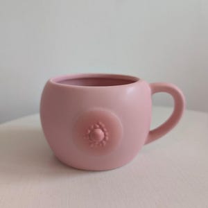 Puede incluir: Una taza de cerámica de color rosa claro mate con una forma redondeada y abombada y una sola asa. La parte frontal de la taza presenta un diseño en relieve circular y elevado en el centro que se asemeja a un pezón rodeado de pequeñas protuberancias.
