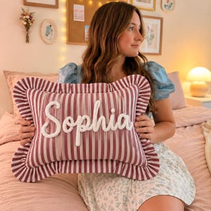May include: A rectangular throw cushion featuring a burgundy and white vertical striped pattern with a ruffled border. The name Sophia is embroidered in white, textured, cursive script across the centre of the cushion front.