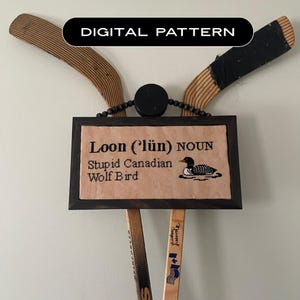 May include: A framed cross-stitch piece with the word "Loon" and a definition, featuring a loon illustration. The frame is mounted with hockey sticks and a puck. The text "DIGITAL PATTERN" is displayed above the frame.