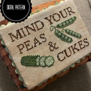 May include: A cross-stitch pattern on beige fabric with green and brown embroidery. The pattern reads "Mind your peas & cukes".